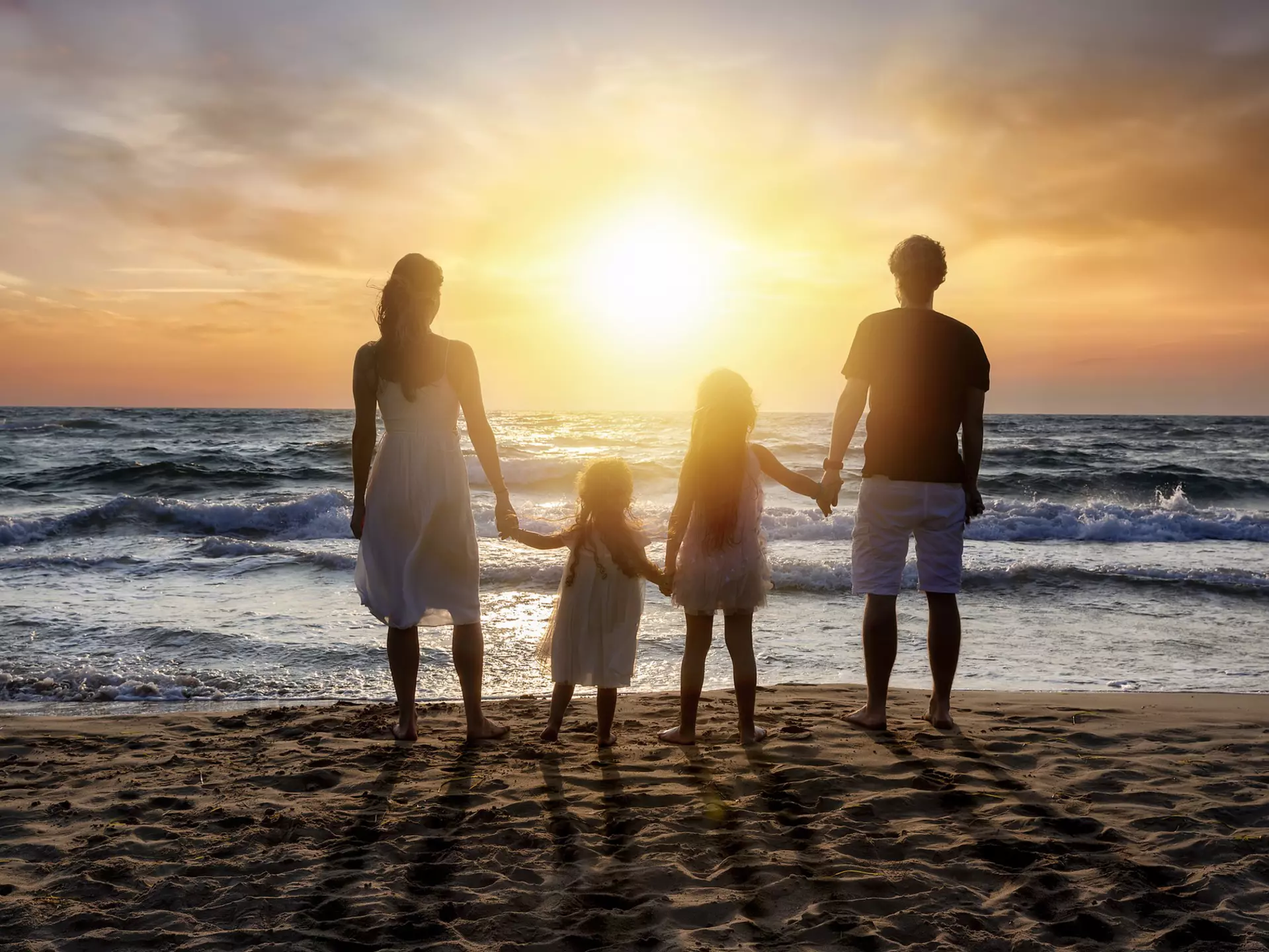 Rear view of parents holding hands with daughters standing at beach against orange sky