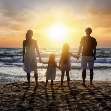 Rear view of parents holding hands with daughters standing at beach against orange sky