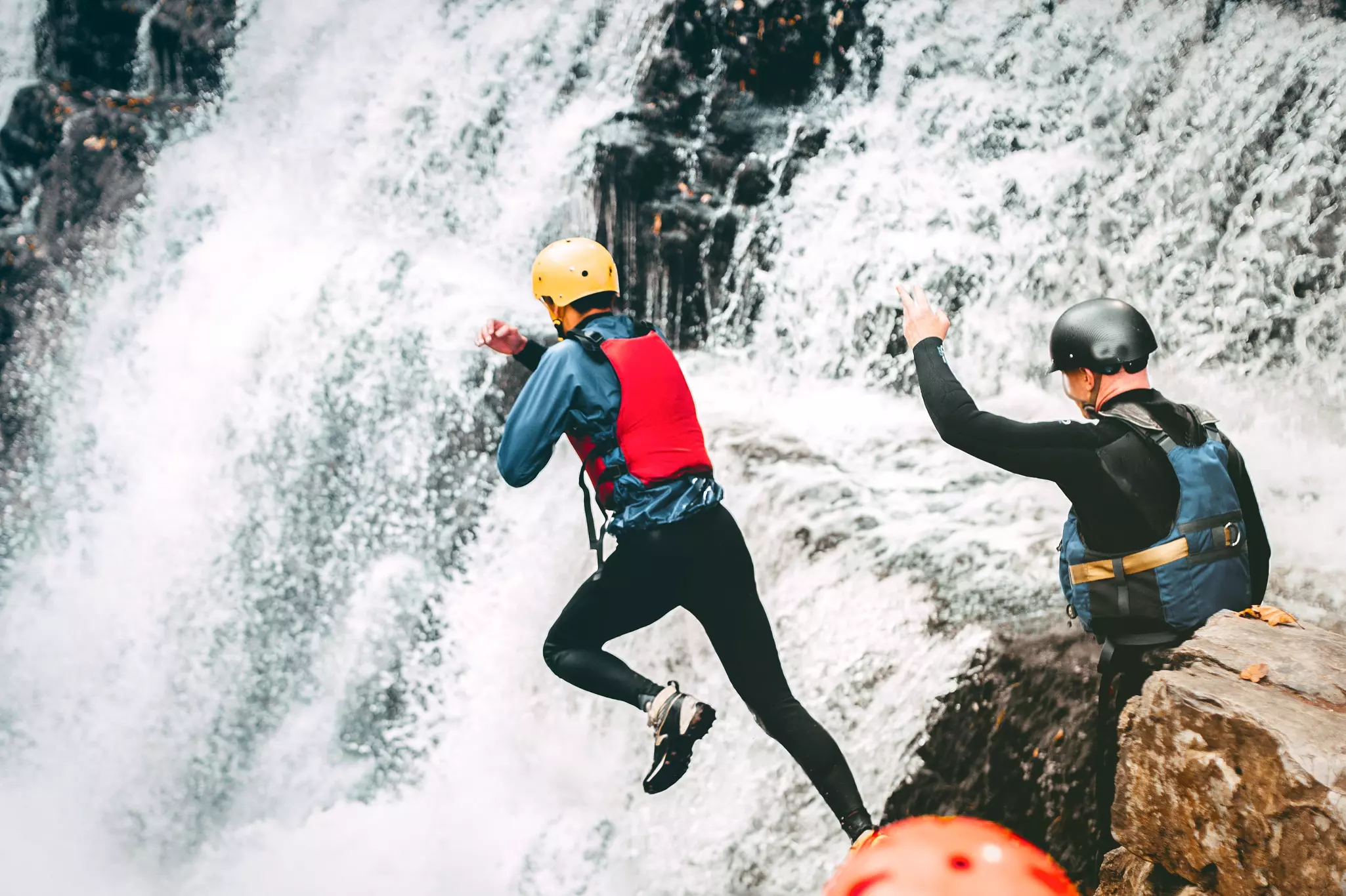 People in wetsuits and helmets jump off a rocky outcrop and into a rushing waterfall.
