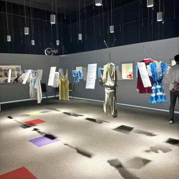 A dim gallery where children's clothes hang from wires beside text panels, with a bicycle and toys in wall cases.