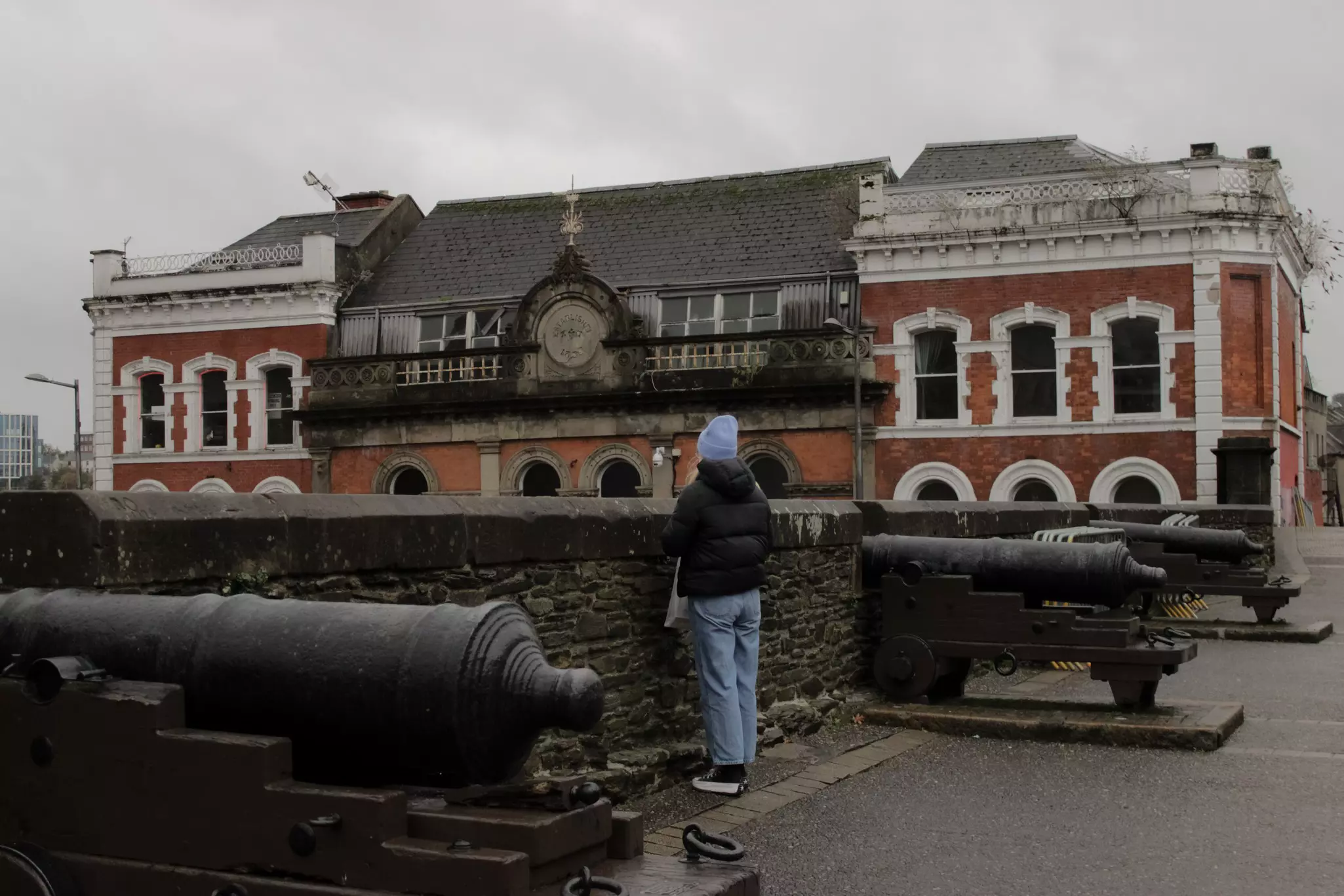 A person wearing a blue hat and blue jeans stands by a stone wall between two cannons.