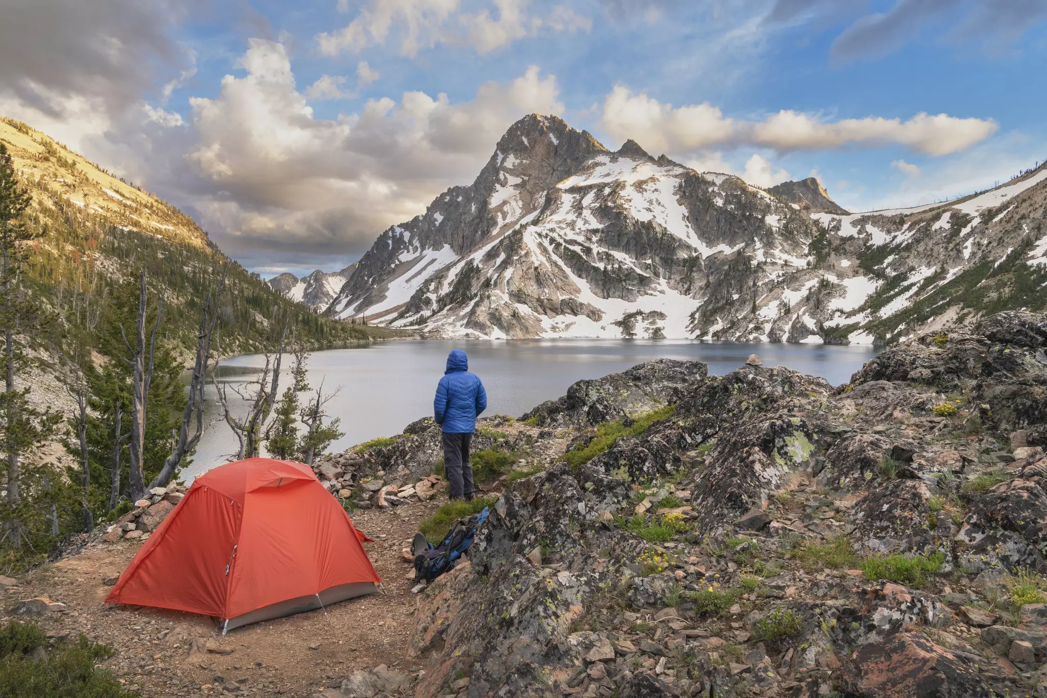 Adult male in blue jacket and red backpacking tent at Sawtooth Lake, Mount Regan is in the distance. Sawtooth Wilderness, Idaho.