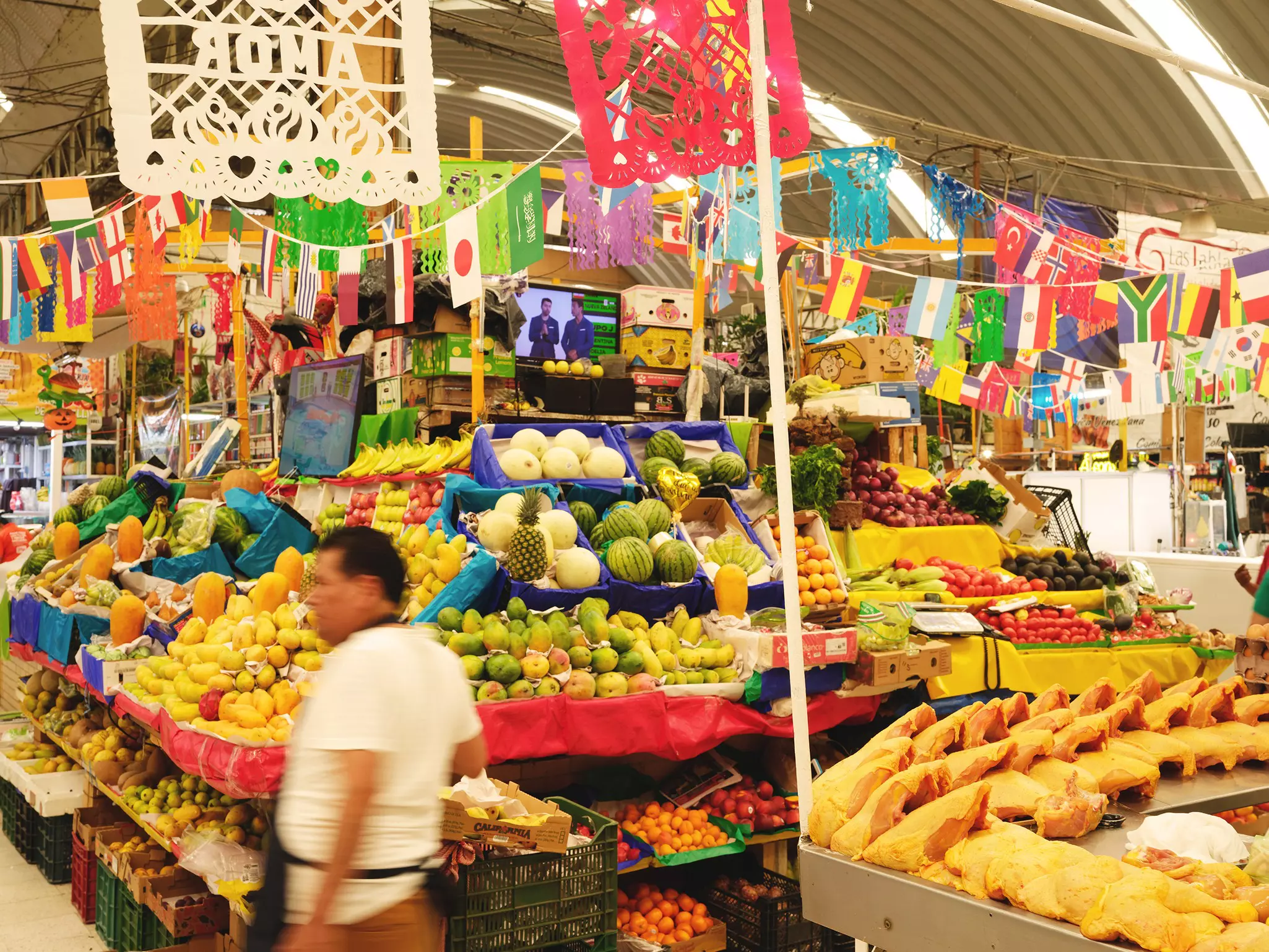A vendor walks past a display of fruits and vegetables inside a market decorated with flags