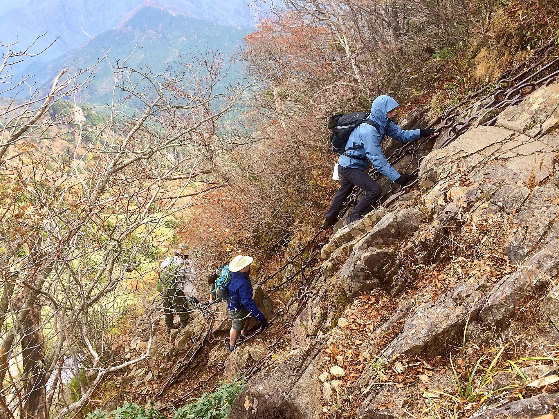 Climbers in raingear with backpacks using chains to pull themselves up a steep, rocky slope with mountains in the distance on an overcast day.