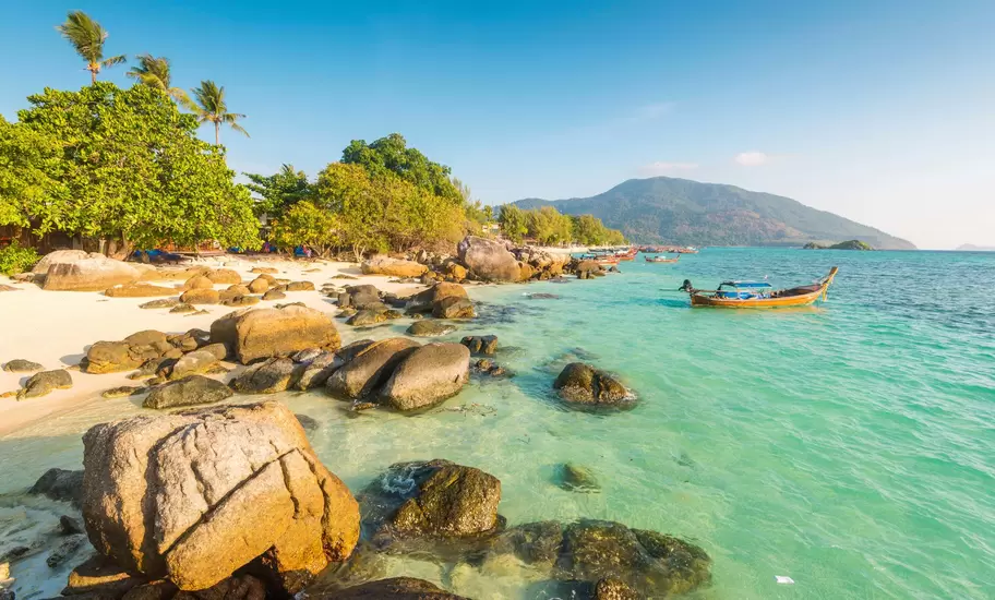 Rounded boulders on a white-sand beach beside turquoise water with a longtail boat moored offshore on Ko Lipe, Thailand.