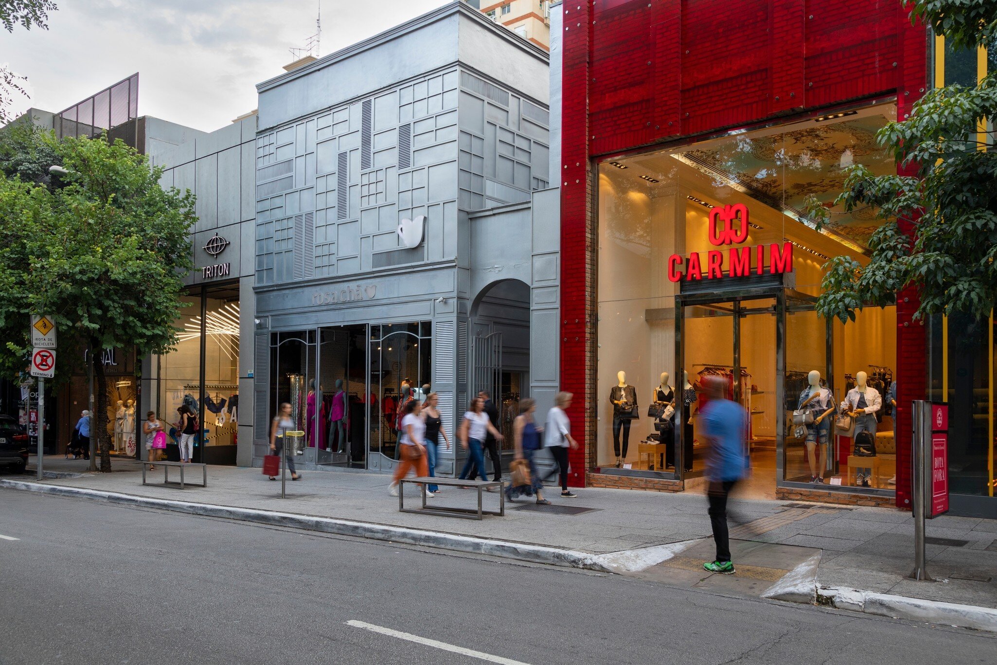 Pedestrians walking along a tree-lined pavement past a grey panelled shopfront and a glossy red store.