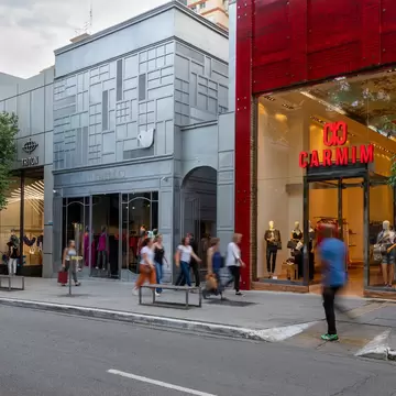 Pedestrians walking along a tree-lined pavement past a grey panelled shopfront and a glossy red store.