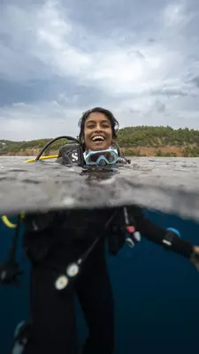 A woman smiles with her head above the water; she has a scuba mask below her chin, and her wetsuit if visible below the water line. 