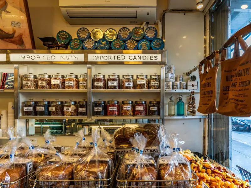 Russ and Daughters, a historic NYC appetizing shop with shelves displaying nuts, candies, and caviar. Baked goods, dried fruits, and branded tote bags are visible.