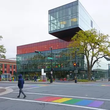 A modern library of stacked glass boxes, the topmost cantilevered out over the street, with a rainbow crossing painted on the road in front.