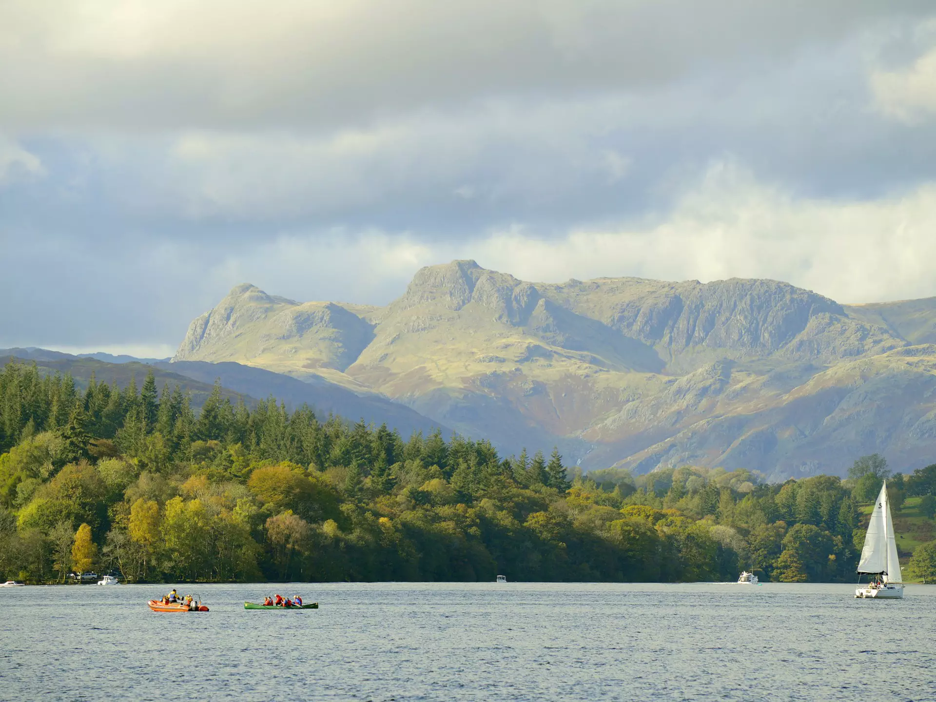 A lake with kayakers out on small boats and a large sail boat with a white sail. It's overlooked by huge green hills in a vibrant national park