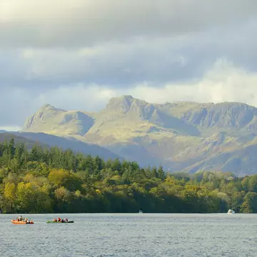 A lake with kayakers out on small boats and a large sail boat with a white sail. It's overlooked by huge green hills in a vibrant national park