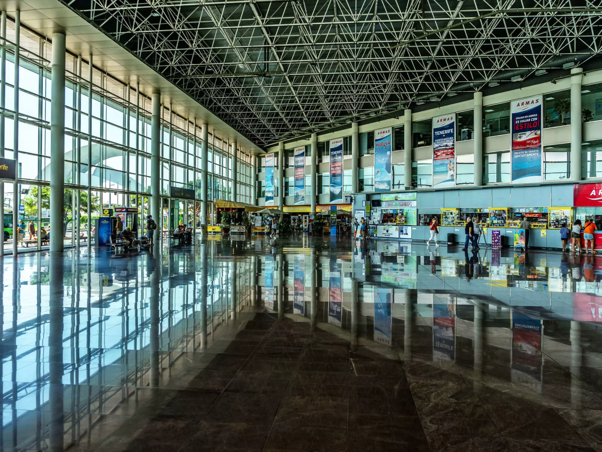 People walking through a bright, modern long-distance bus station with a wall of windows on one side