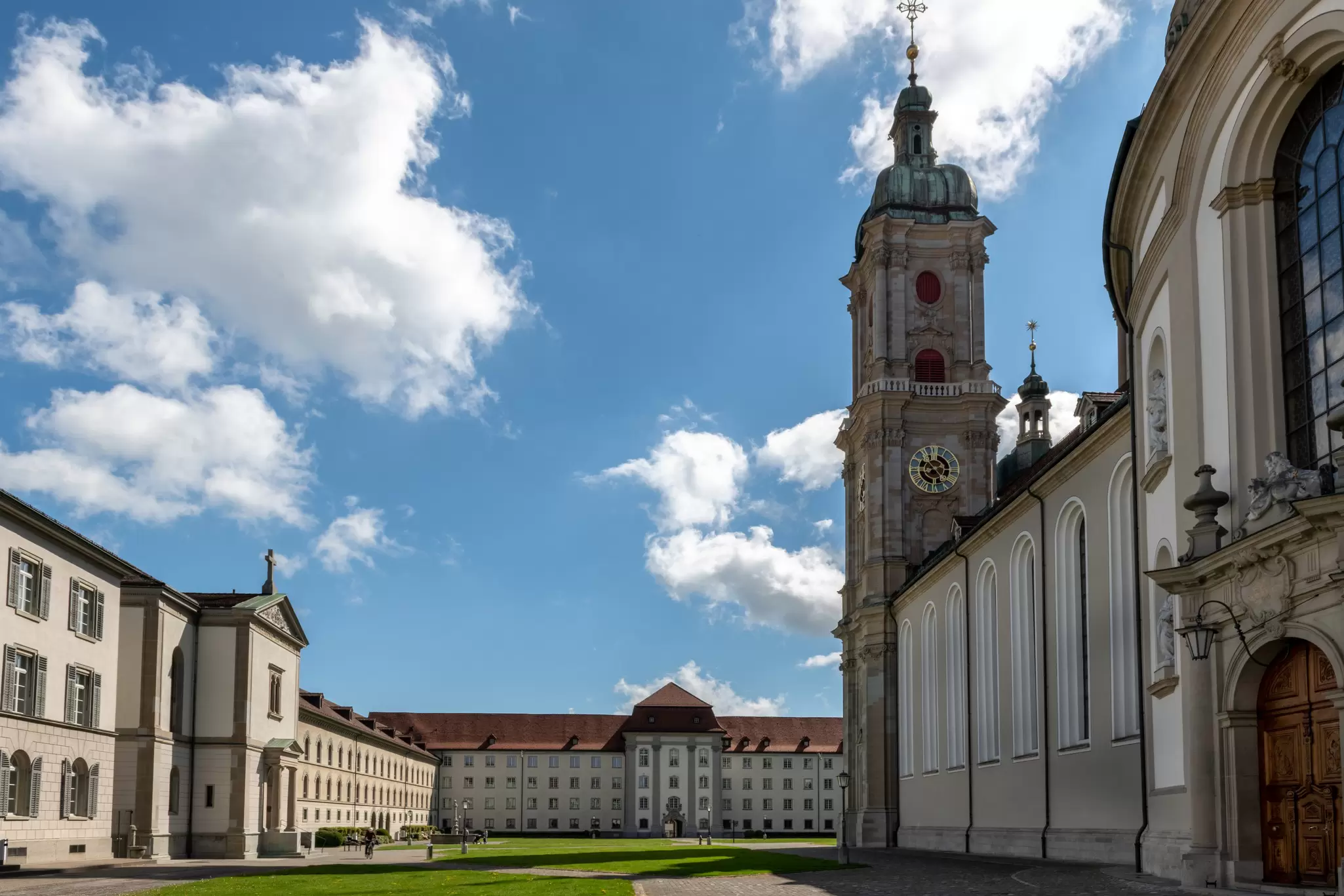 Exterior of the Abbey of St Gall. A high white wall extends into the distance, where the grand tower of a cathedral is visible.