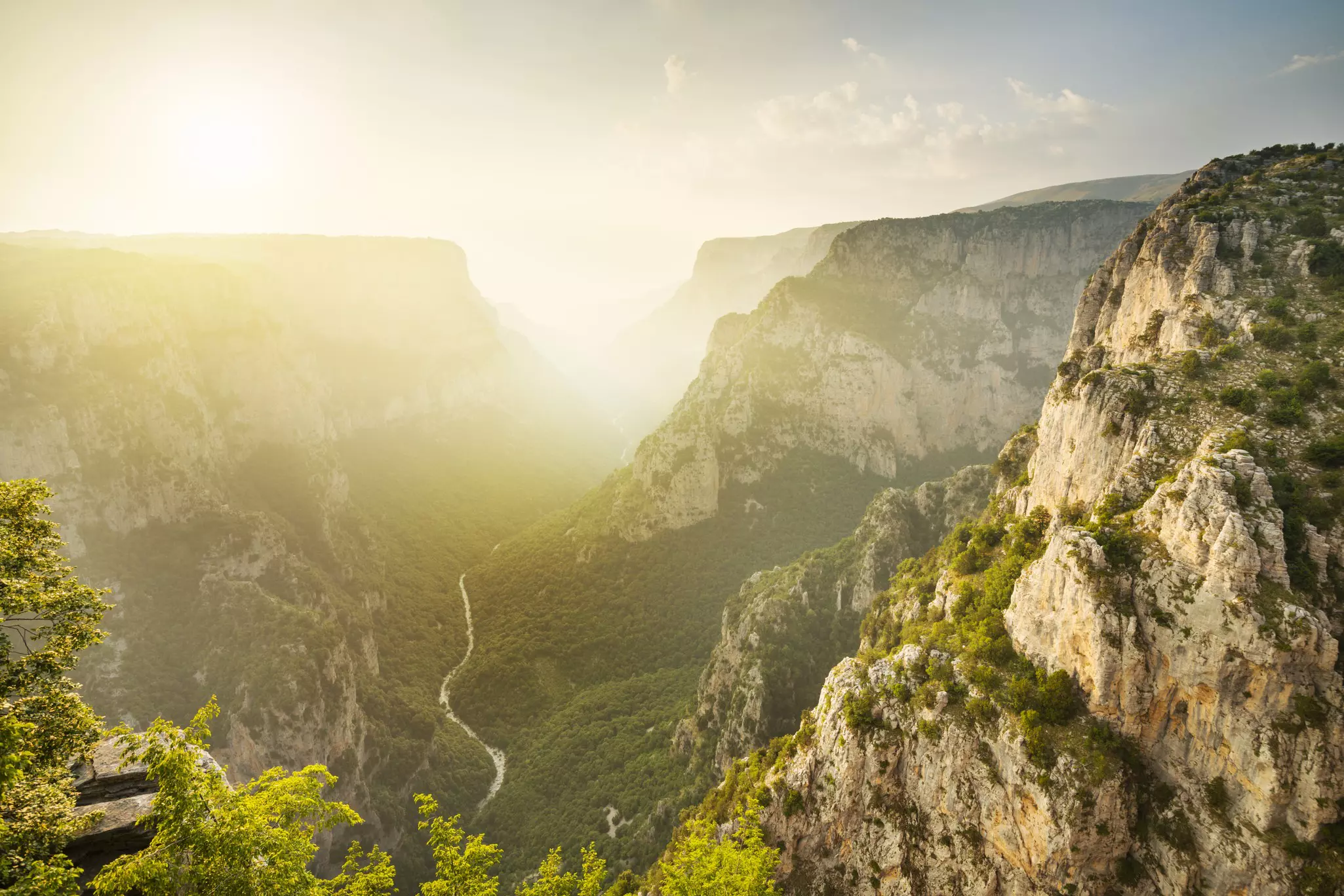 Sunlight over a deep gorge. A river runs through it while trees grow on the rocky mountains at either side.