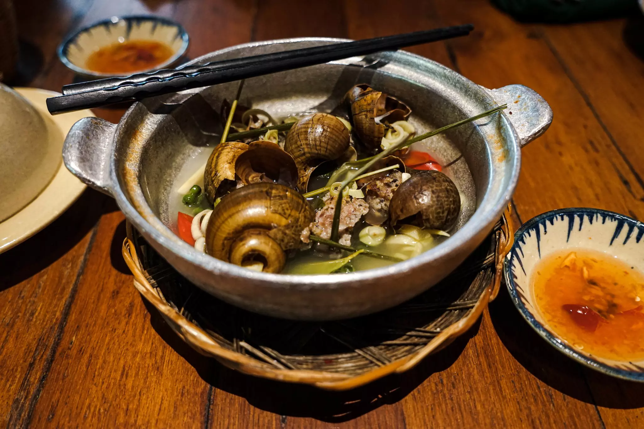 Sea snails in broth sit in a blue ceramic bowl placed on a wooden table at a restaurant.