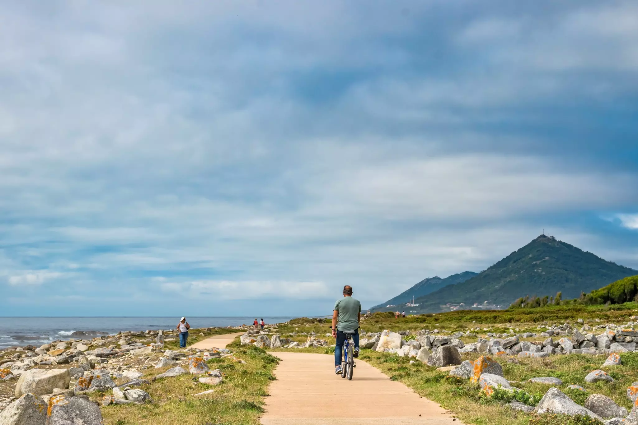 A cyclist on a path near the sea, with rocks on either side of the path and a steep hill in the distance.
