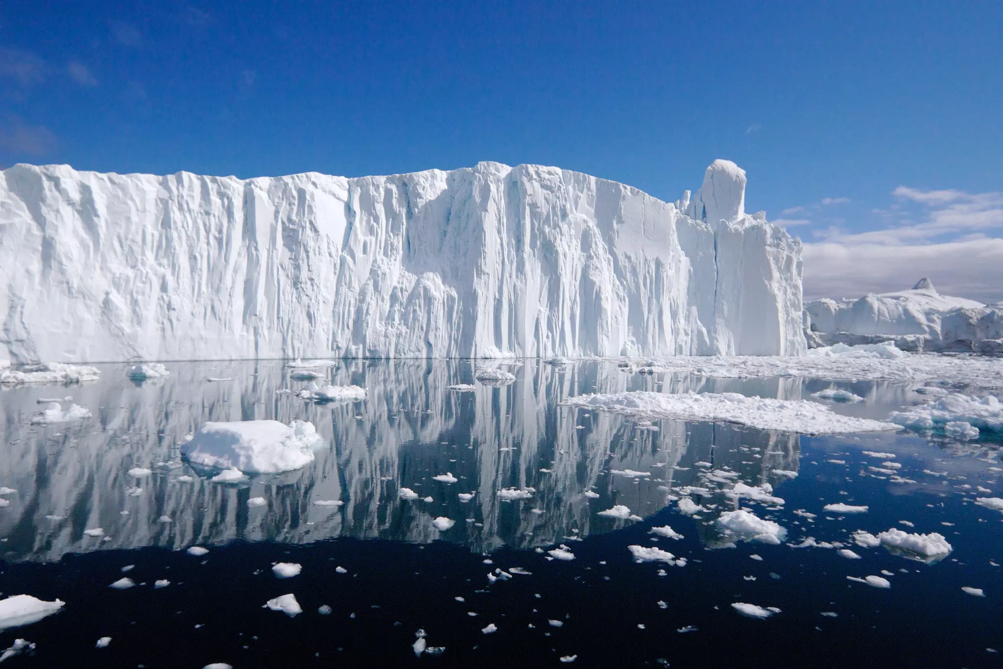 The edge of a glacier forming a vast cliff of bright white ice against an ocean dotted with smaller pieces of ice.