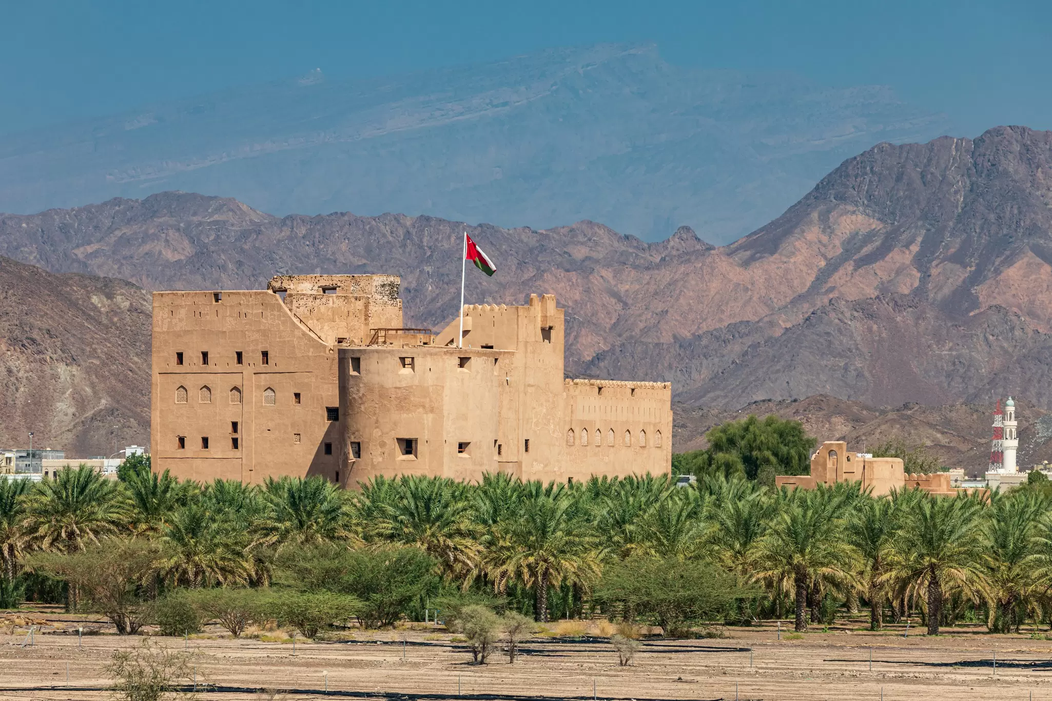 A large stone ancient fortress stands between palm trees and a mountain range; Jabreen Castle in Bahla, Oman