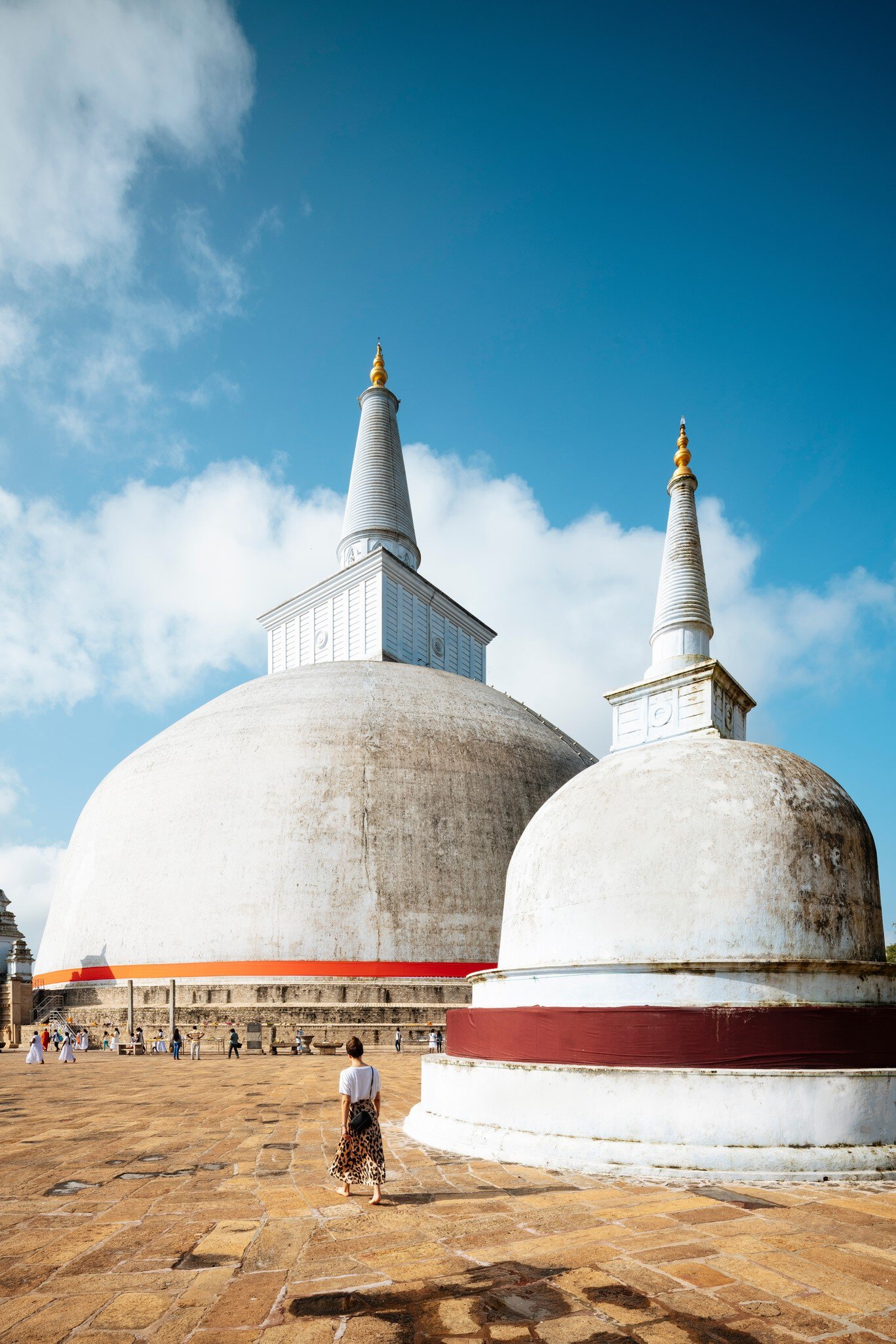 Two white dome-shaped stupas with tall spires on a stone terrace, a woman and small groups of people walking nearby