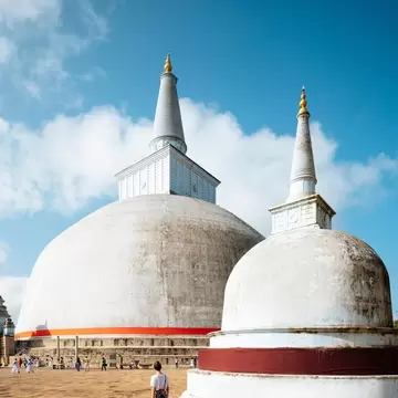 Two white dome-shaped stupas with tall spires on a stone terrace, a woman and small groups of people walking nearby