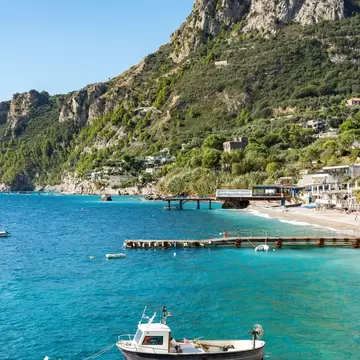 Small white-cabin boat moored in clear blue water off Massa Lubrense, with a pebble beach and a steep limestone peak behind