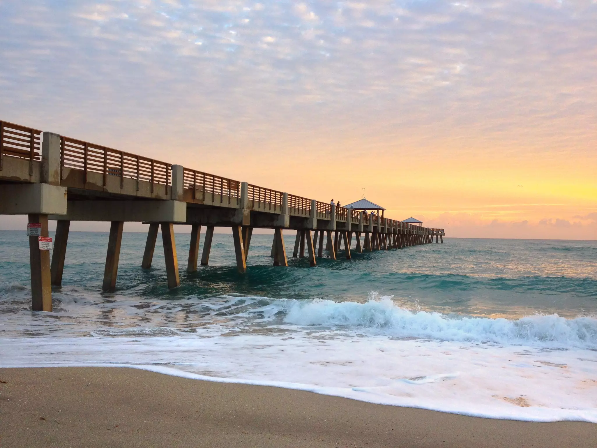 A long pier over soft waves crashing on a sandy beach at sunrise.