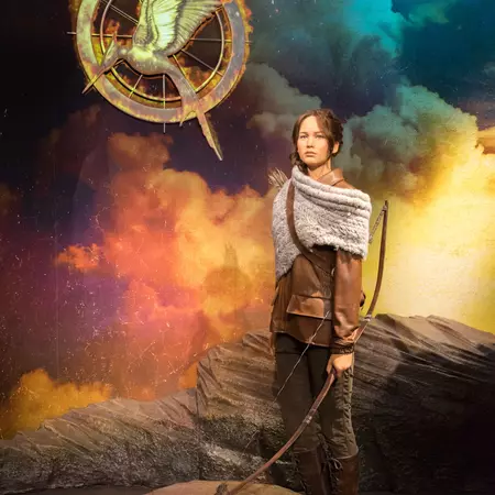 Wax figure of an actress as a movie character dressed in brown, holding a bow with a symbol behind her