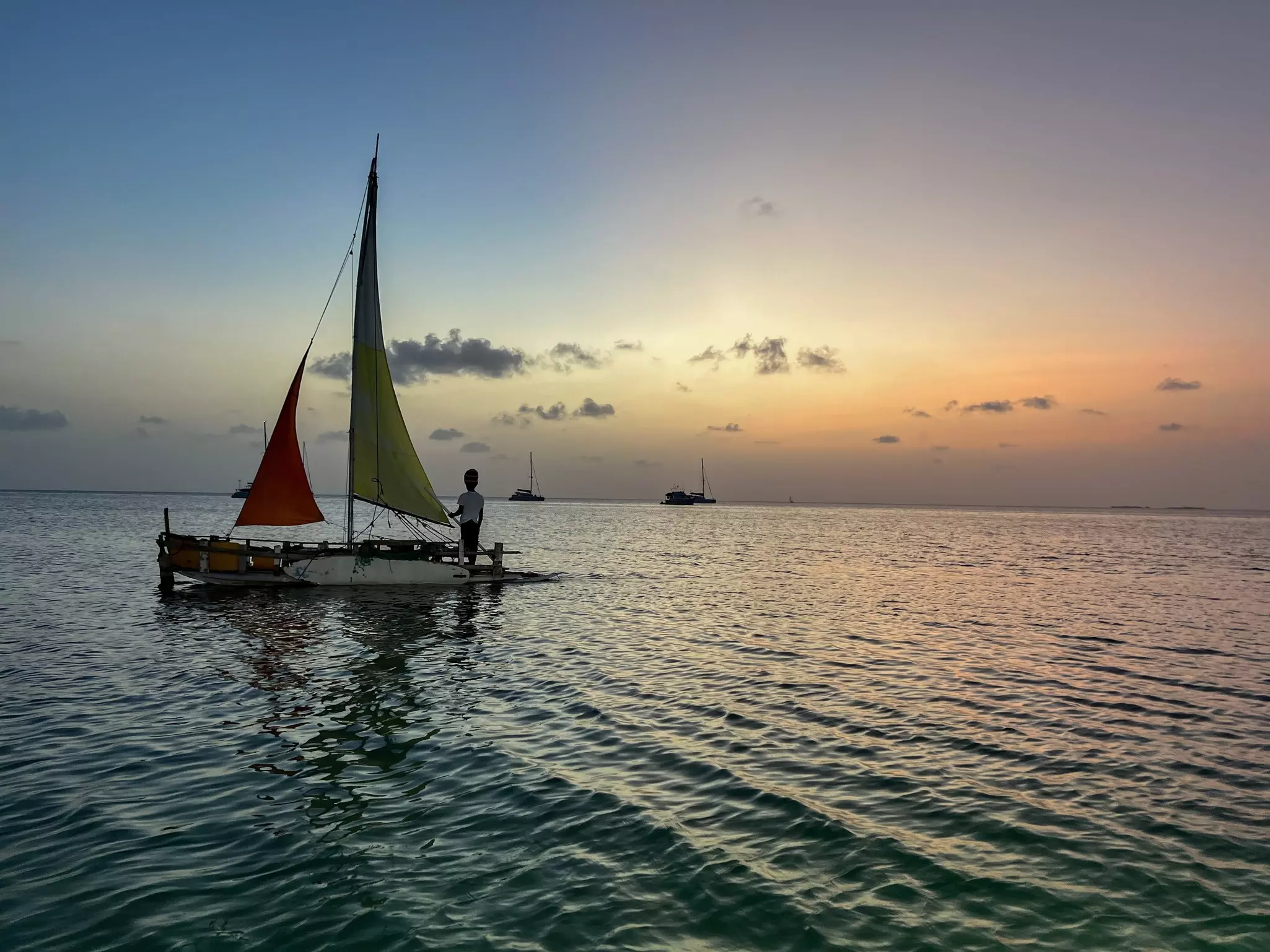 A person stands on a small boat with one red and one yellow sail at sunset.