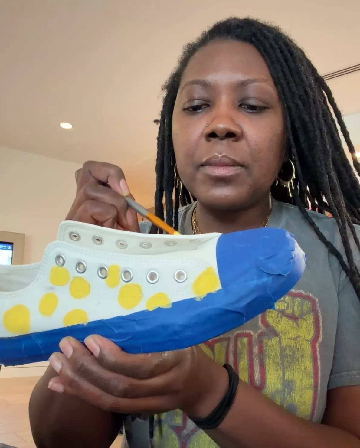 A Black woman with locks paints yellow dots on a pair of white sneakers.