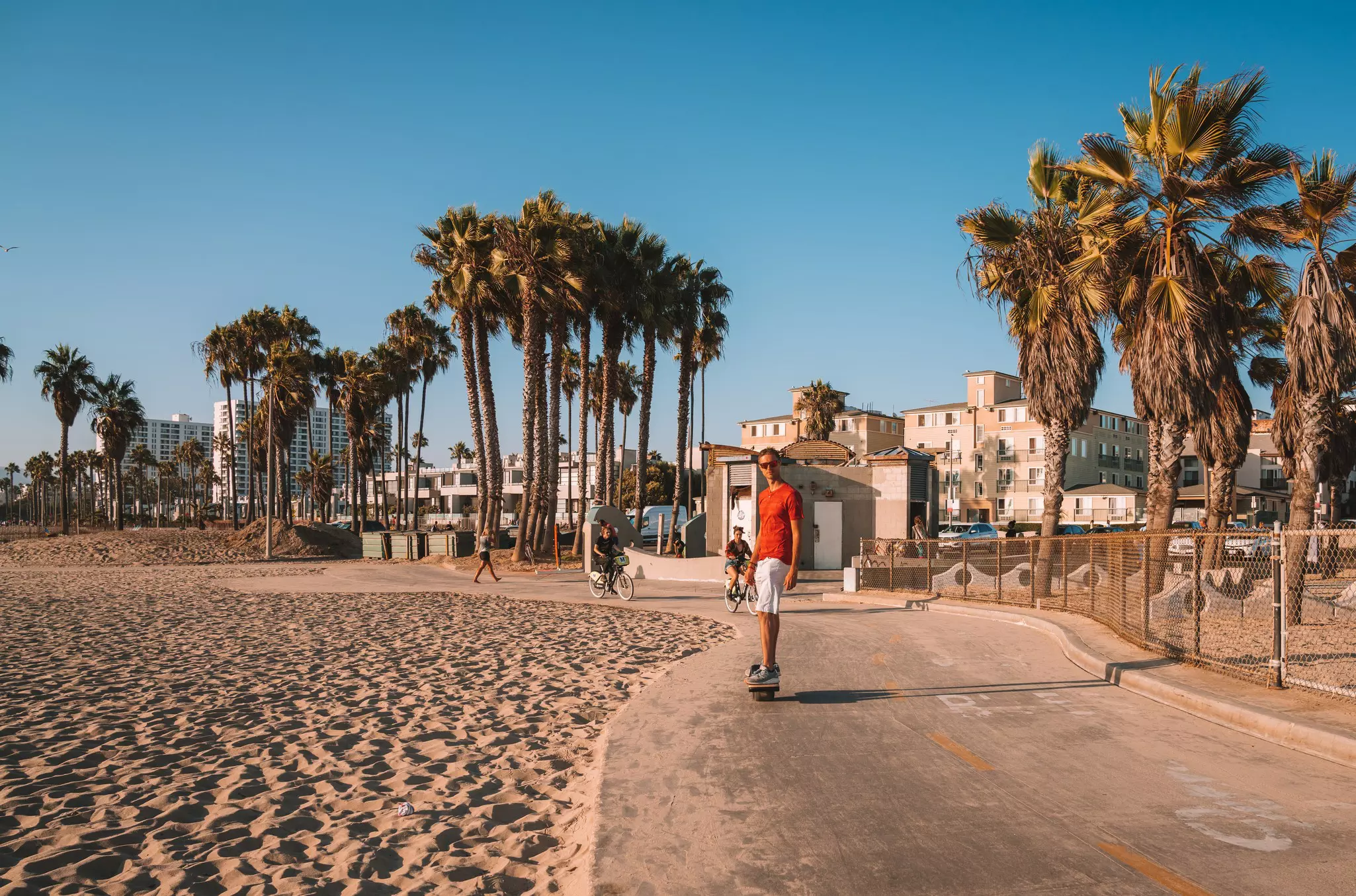 Young man riding a one wheeler down a paved path near a beach surrounded by palm trees