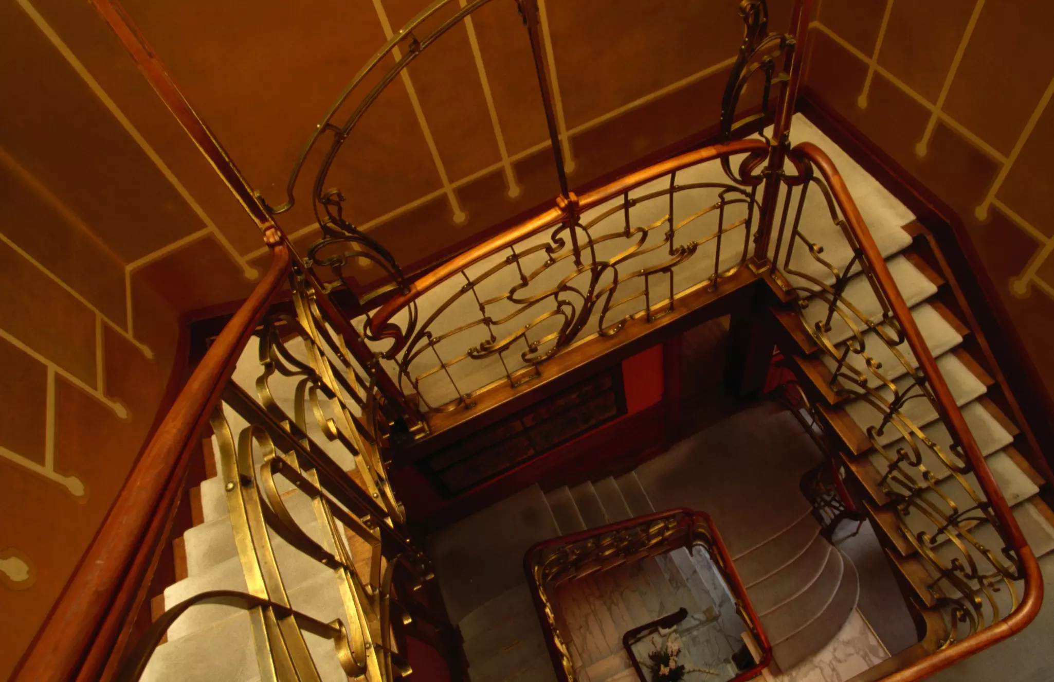 A staircase with sinuous metalwork and rich wood paneling is seen from above, with other floors of a house visible through the well.