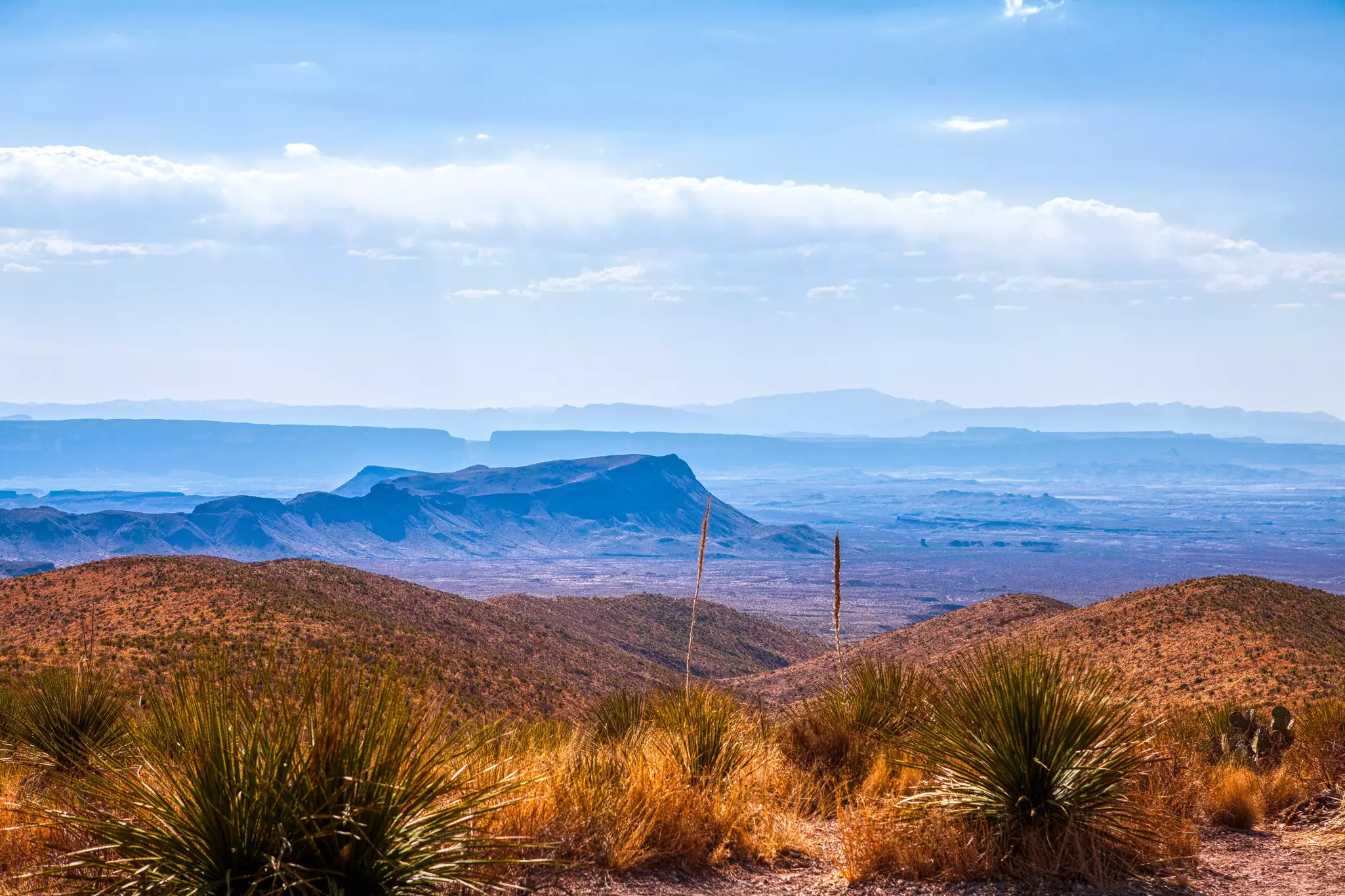 A view from a hill of of a rocky outcrop in a desert landscape. The landscape appears blue in a hazy effect.