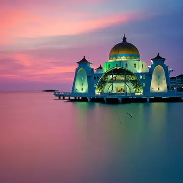 Gold-domed Straits Mosque standing on stilts above calm water in Melaka at sunset, with its white minaret beside it