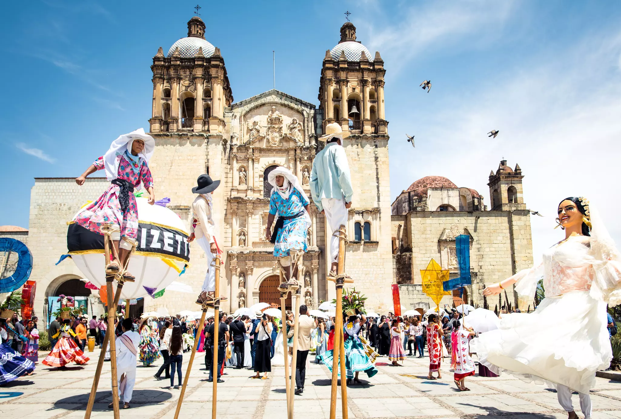 People walk on stilts and dance in papier-mâché masks on a plaza in front of a baroque church.