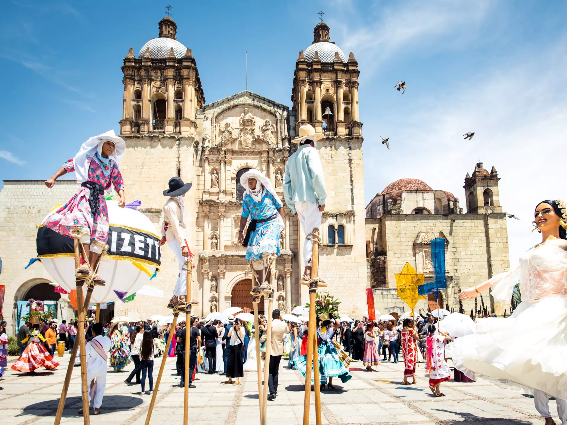 People walk on stilts and dance in papier-mâché masks on a plaza in front of a baroque church.