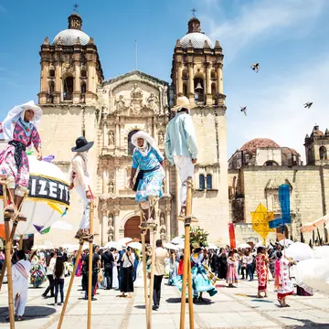 People walk on stilts and dance in papier-mâché masks on a plaza in front of a baroque church.