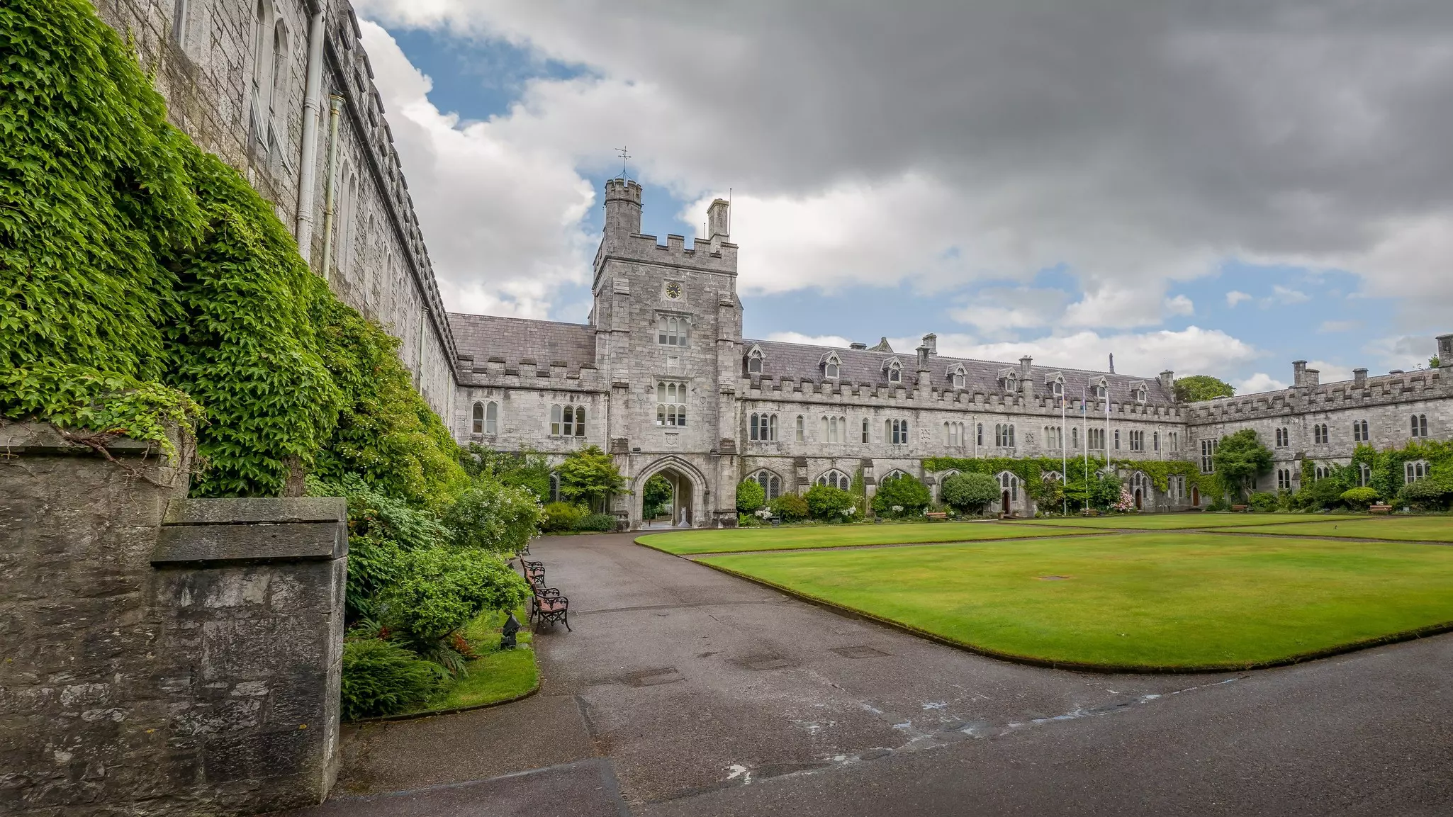 A wide view of a two-story gray stone building with gables and a tower at one end; a grass quadrangle is in front of the building.