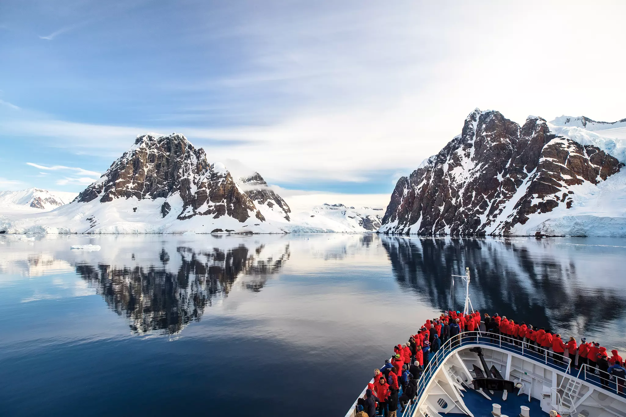 Passengers aboard an expedition ship gather around the bow to look at the surrounding mountains covered in snow in an antarctic ocean.