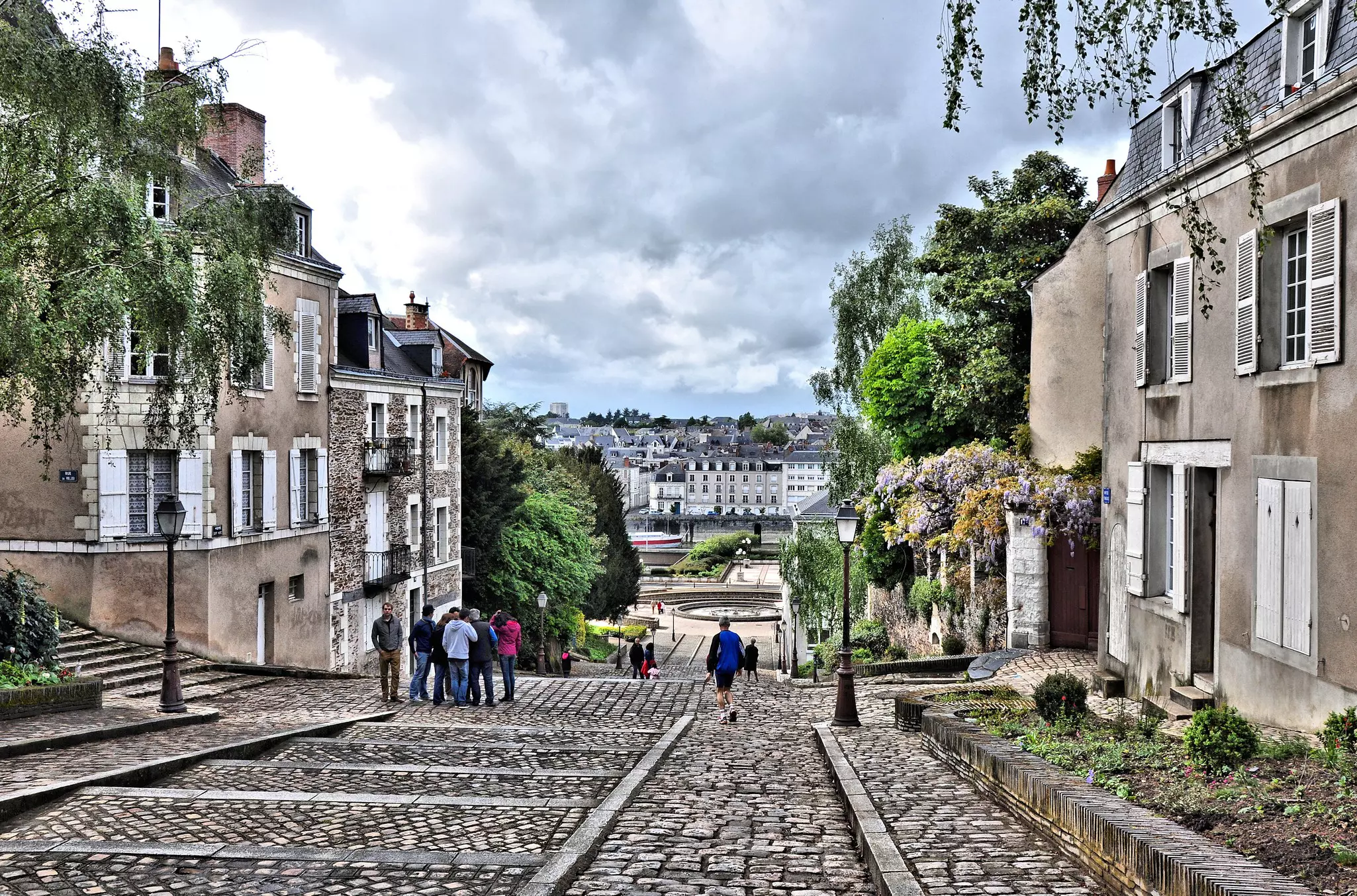 People are seen walking on a sloped, wide cobbled street in a city.