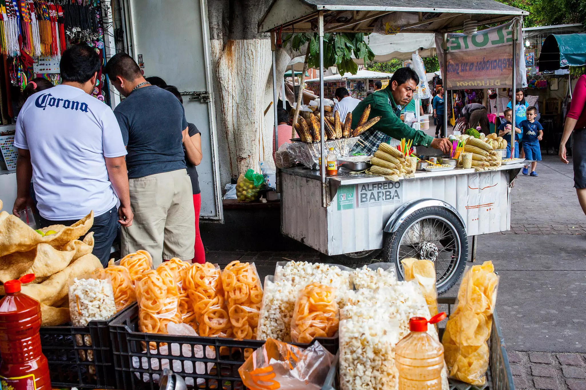 A man selling corn at a street stall. Other food items are in crates in the foreground.