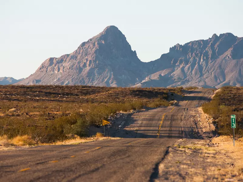 A two-lane road winds through low green bush with large rocky mountains in the background.