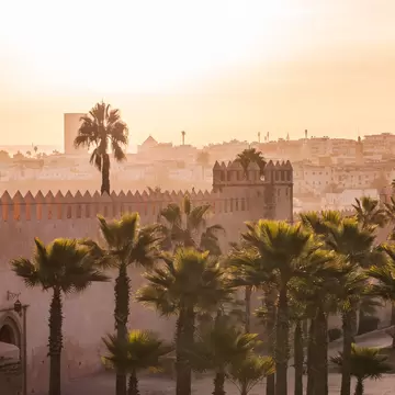 Lamps on a plaza by ancient stone walls and towers; a city with white buildings is above on a hillside.