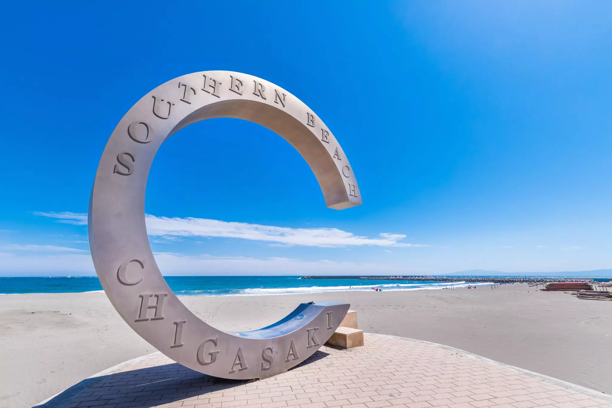 A stone sculpture depicting the letter C stands on a beach under a blue sky.
