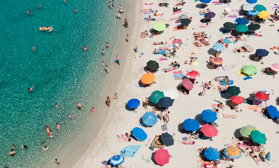 Overhead view of a busy pale-sand beach in Tropea with colourful parasols and sunbathers beside clear turquoise water