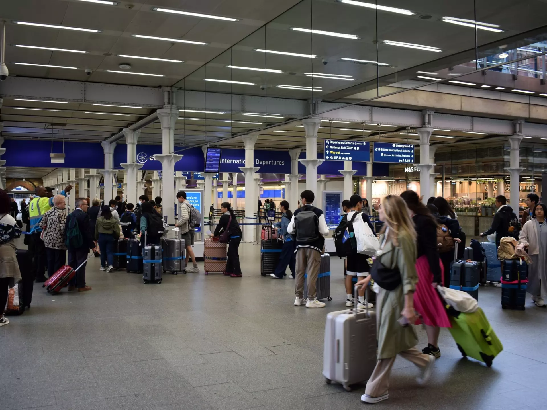 Passengers with luggage queue by check-in counters at a train station.