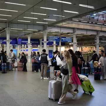 Passengers with luggage queue by check-in counters at a train station.