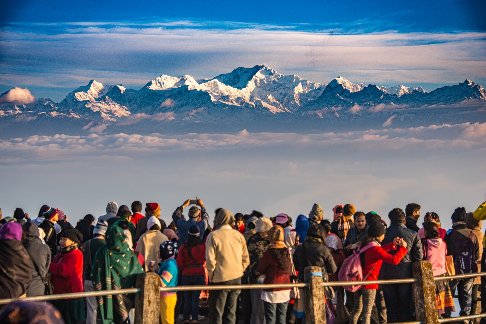 A large group of people gather at a viewpoint to watch the sunrise over a snow-capped mountain.