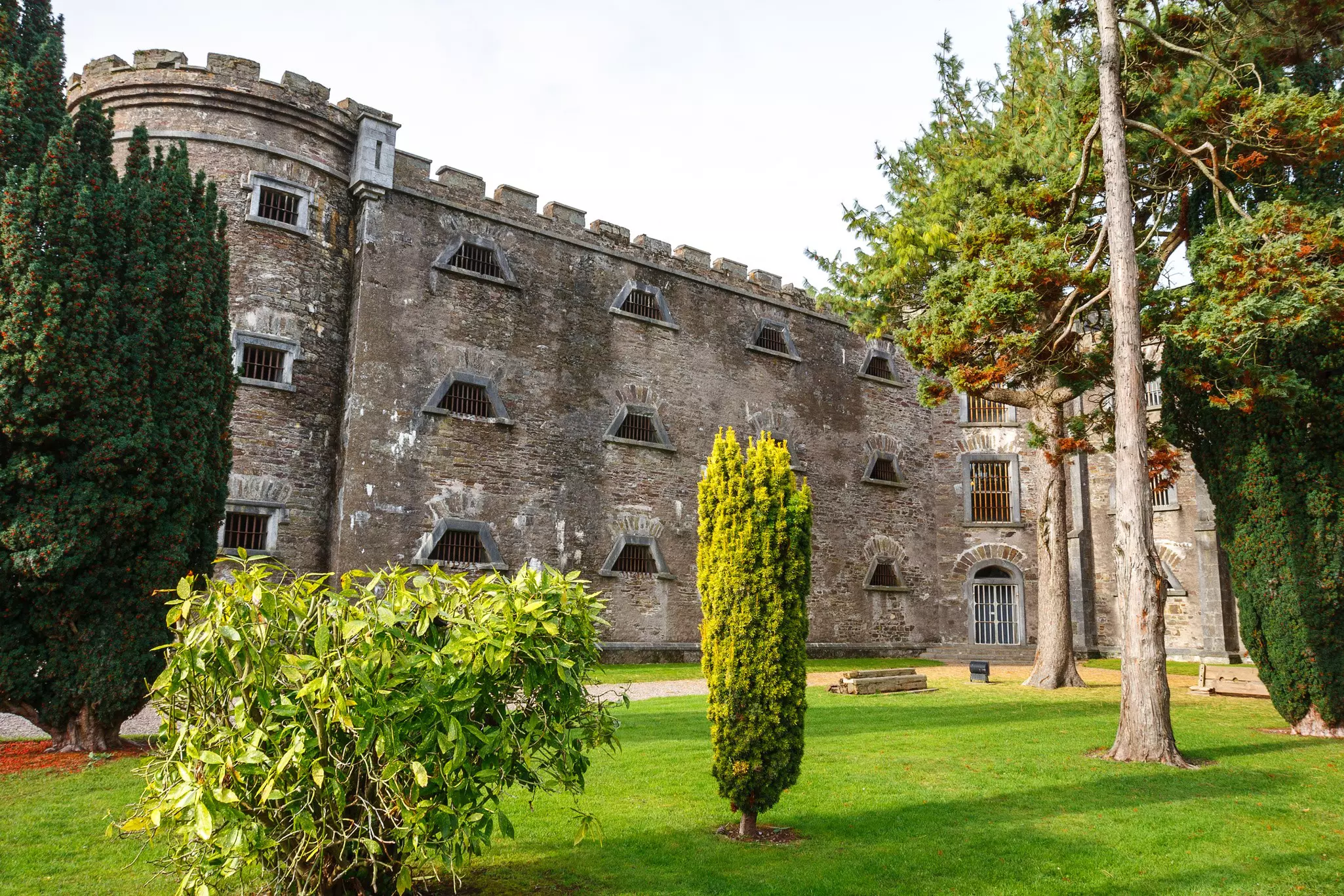 A lawn with bushes and trees in the foreground leads to the high stone walls of a historic jail in Cork city, Ireland.