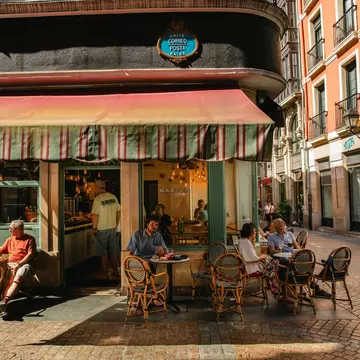 People sit on a sidewalk outside a restaurant with a striped awning in the city of Bilbao, Spain